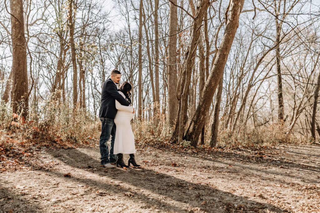 Maternity session at flowing well park where couple is posed together in front of wooded trees